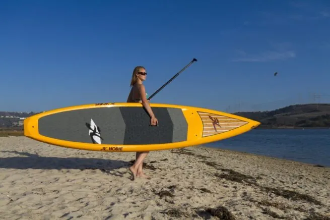 Woman standing on the shore holding ayellow Hobie Elite touring paddleboard.