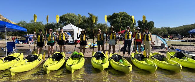 Bachelorette party by yellow kayaks on lakeshore. Riverbound Sports Paddle Company